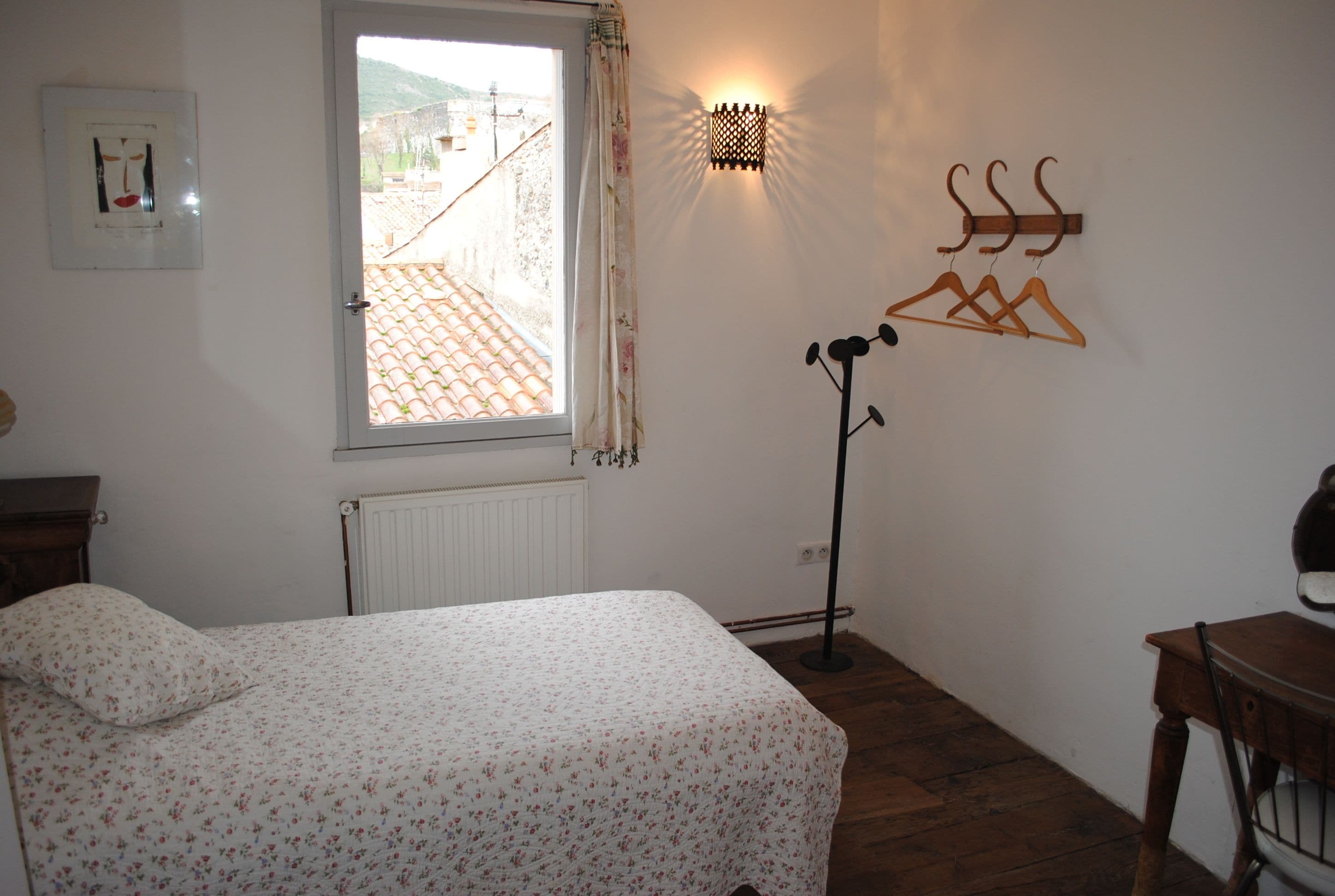 Un dormitorio con cama, un radiador, una ventana con vistas, una lámpara de pared y un perchero.