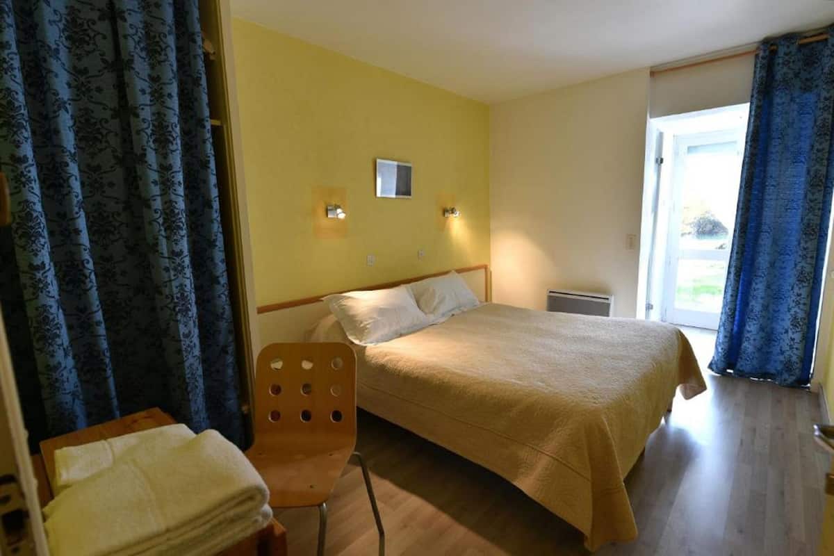 A bedroom with a bed, a chair, a small table with a towel, and a window with curtains.