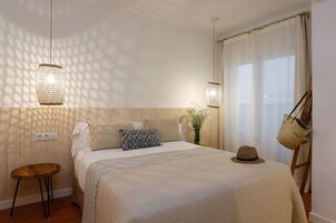 Un dormitorio con cama, lámparas de noche, un taburete de madera, una cesta y una ventana con cortinas.