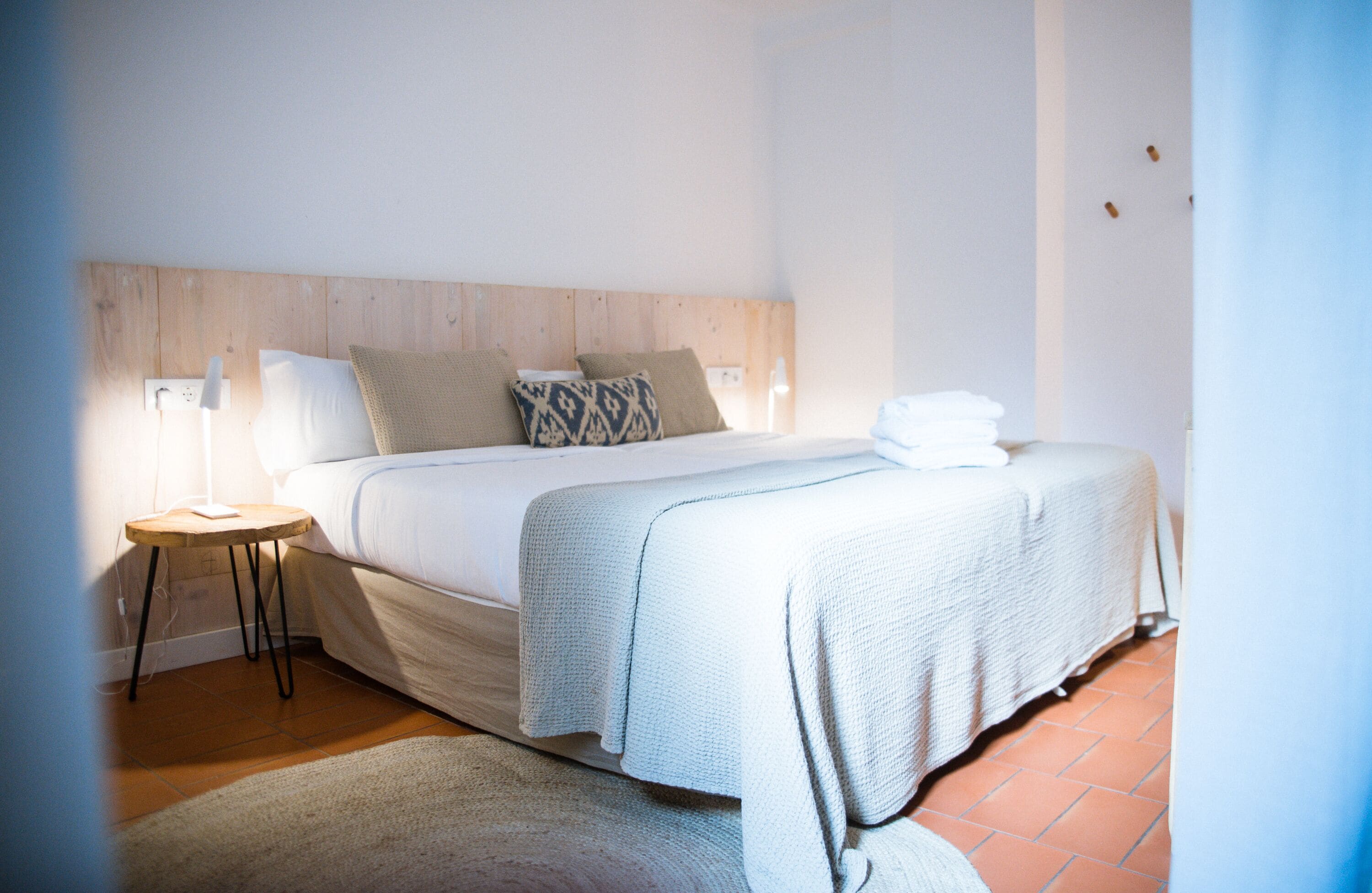 Un lit bien fait, avec une tête de lit en bois, une petite table de chevet sur laquelle se trouve une lampe, et une serviette blanche posée sur le lit.