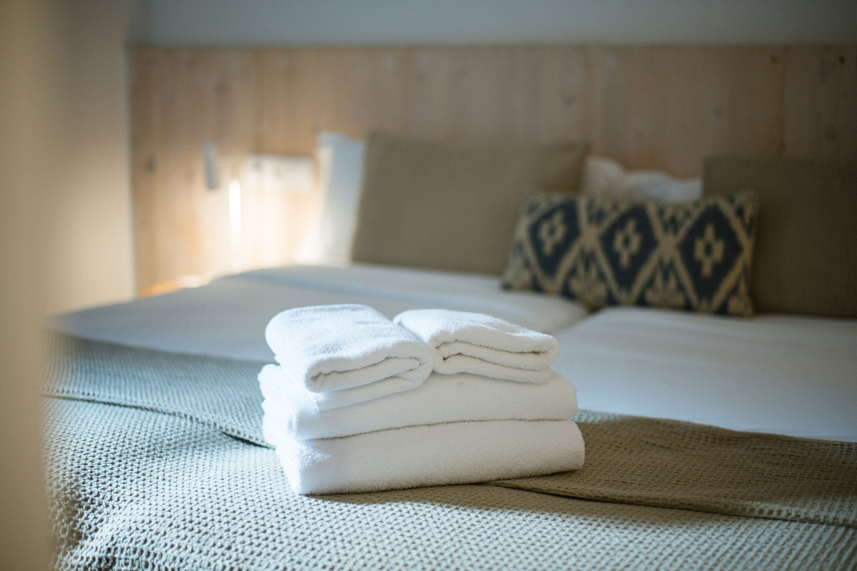 A neatly made bed with a folded white towel on a textured bedspread, a wooden headboard, and patterned pillows.