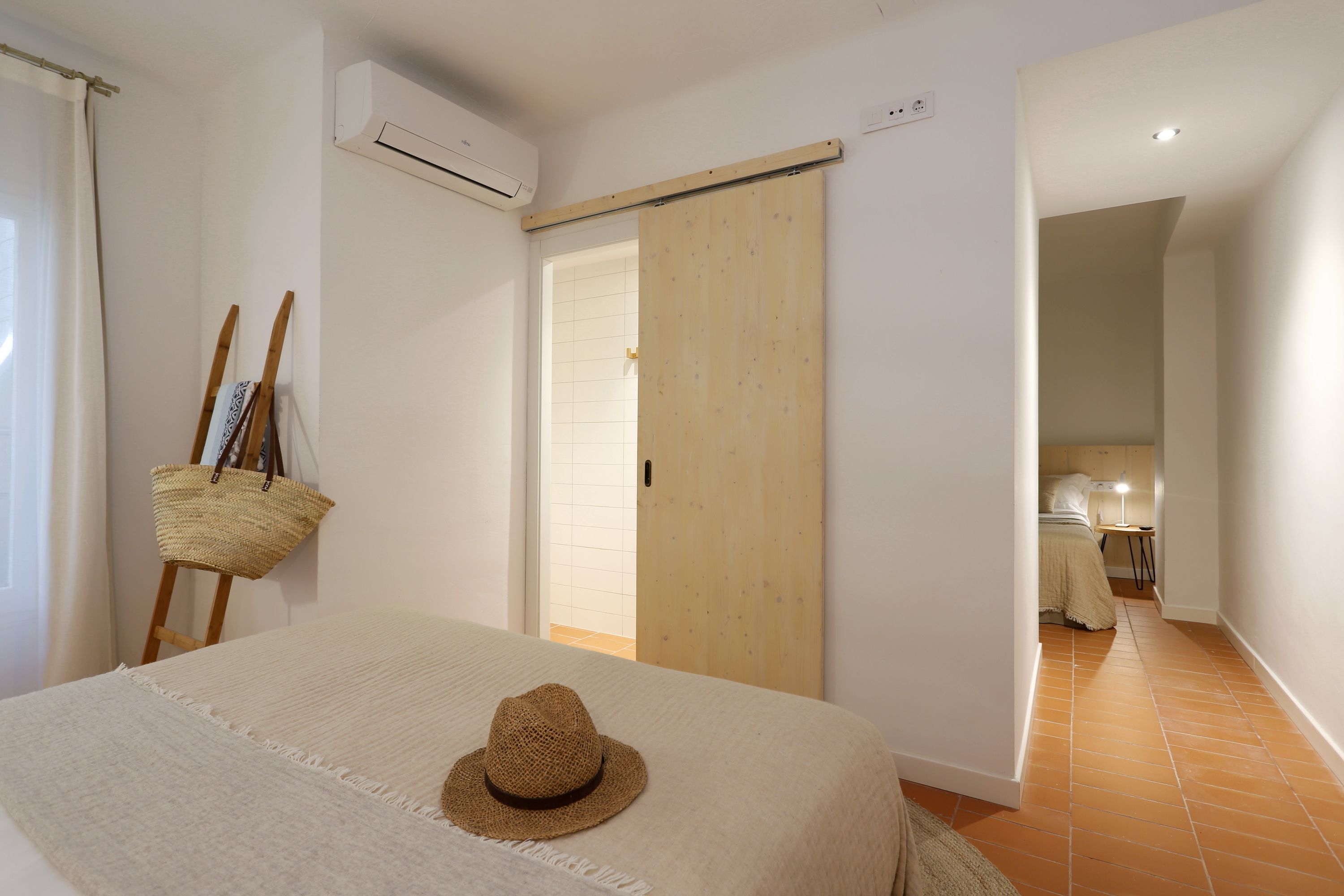 Un dormitorio con una cama, un sombrero de paja sobre la cama, una puerta de madera y un pasillo que conduce a otra habitación.
