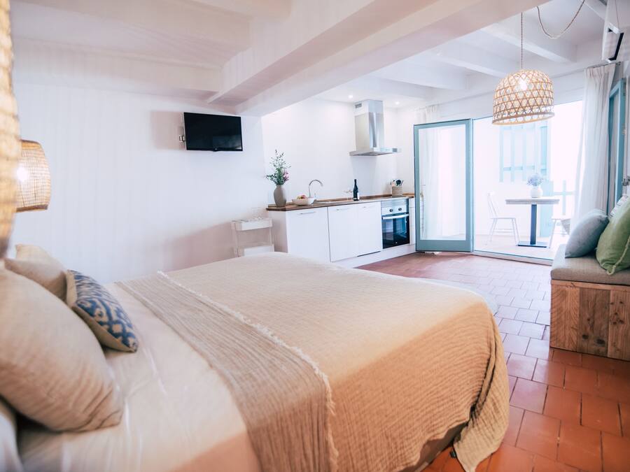 Un dormitorio con una cama grande, un banco de madera con cojines y vistas a la cocina y al balcón.