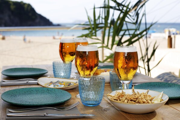 A table set with plates, cutlery, and glasses of beer on a wooden table with a beach view in the background. - Hostal Ses Negres (Begur)
