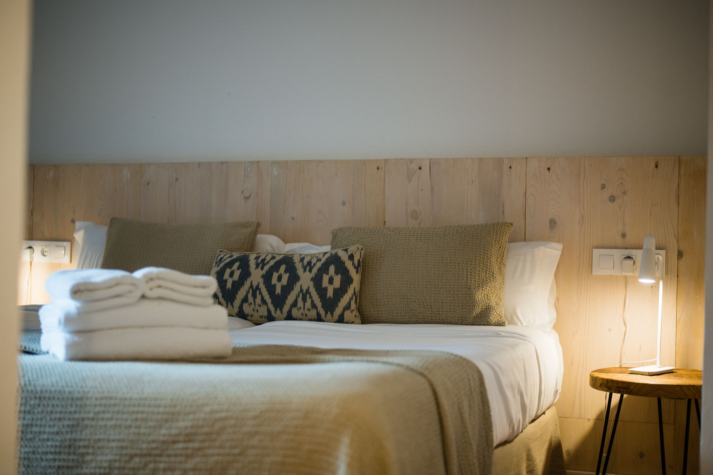 A neatly made bed with folded towels, a patterned pillow, and a bedside lamp on a wooden nightstand.