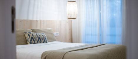 A neatly made bed with a beige blanket and two pillows, a bedside lamp, and a window with sheer curtains.