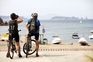 Dos ciclistas en una pasarela de madera junto al mar, con barcos al fondo.