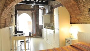 A room with a brick archway, a bed with a colorful blanket, a desk with a chair, a small table with a lamp, and a kitchenette with cabinets and a window. - Vico Del Poeta (Assisi)