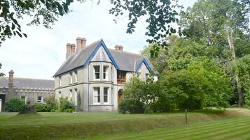Stately building with a gabled roof and symmetrical facade, set amidst a well-manicured lawn and lush trees.