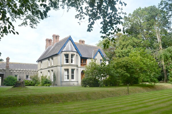 Une grande maison historique, dotée d’une tour d’angle imposante et d’une décoration bleue, entourée de pelouses bien entretenues et d’arbres majestueux.
