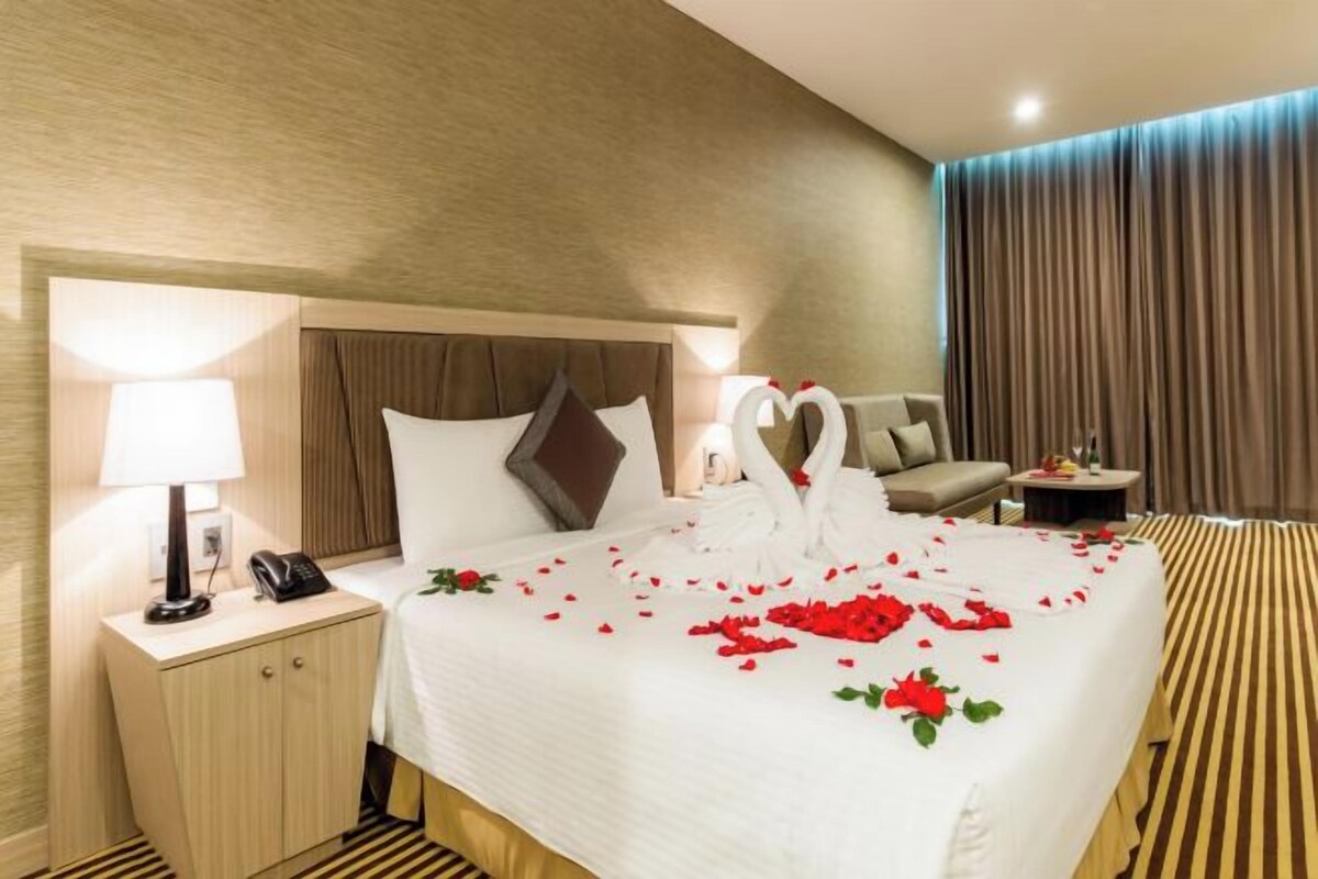 A hotel room with a neatly made bed, a bedside lamp, a telephone, a sofa, and a small table with a vase.