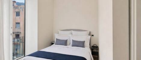 A bedroom with a bed, a nightstand, a window, and a view of buildings outside.