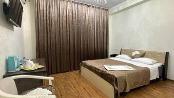 A hotel room with a bed, a desk with a chair, a television, a chandelier, and a large window with curtains.