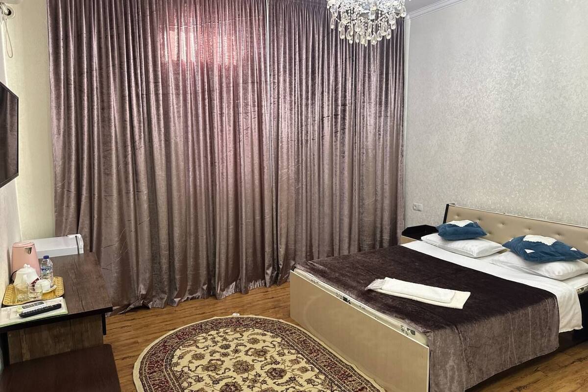 Une chambre d’hôtel avec un lit, un bureau et une chaise, un lustre et une grande fenêtre avec des rideaux.
