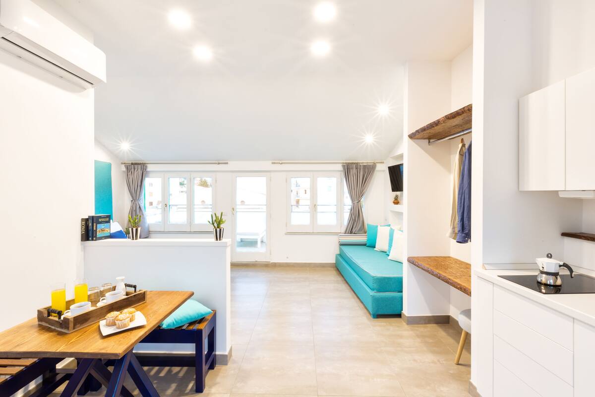 Una cocina moderna con encimera de madera, un fregadero y una estufa. Hay un comedor con mesa y sillas, y una zona de estar con sofá y cojines.