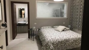 A bedroom with a bed, a bedside table, a mirror, a window with a curtain, and a bathroom with a sink and mirror. - B&B Geolja (Gioia Tauro)