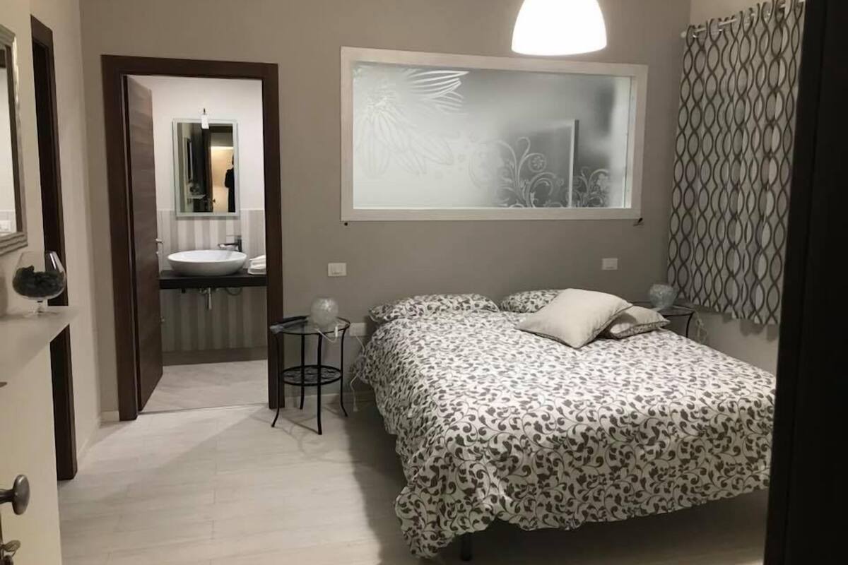 Quarto com cama, mesa de cabeceira, espelho, janela com cortina e banheiro com pia e espelho.