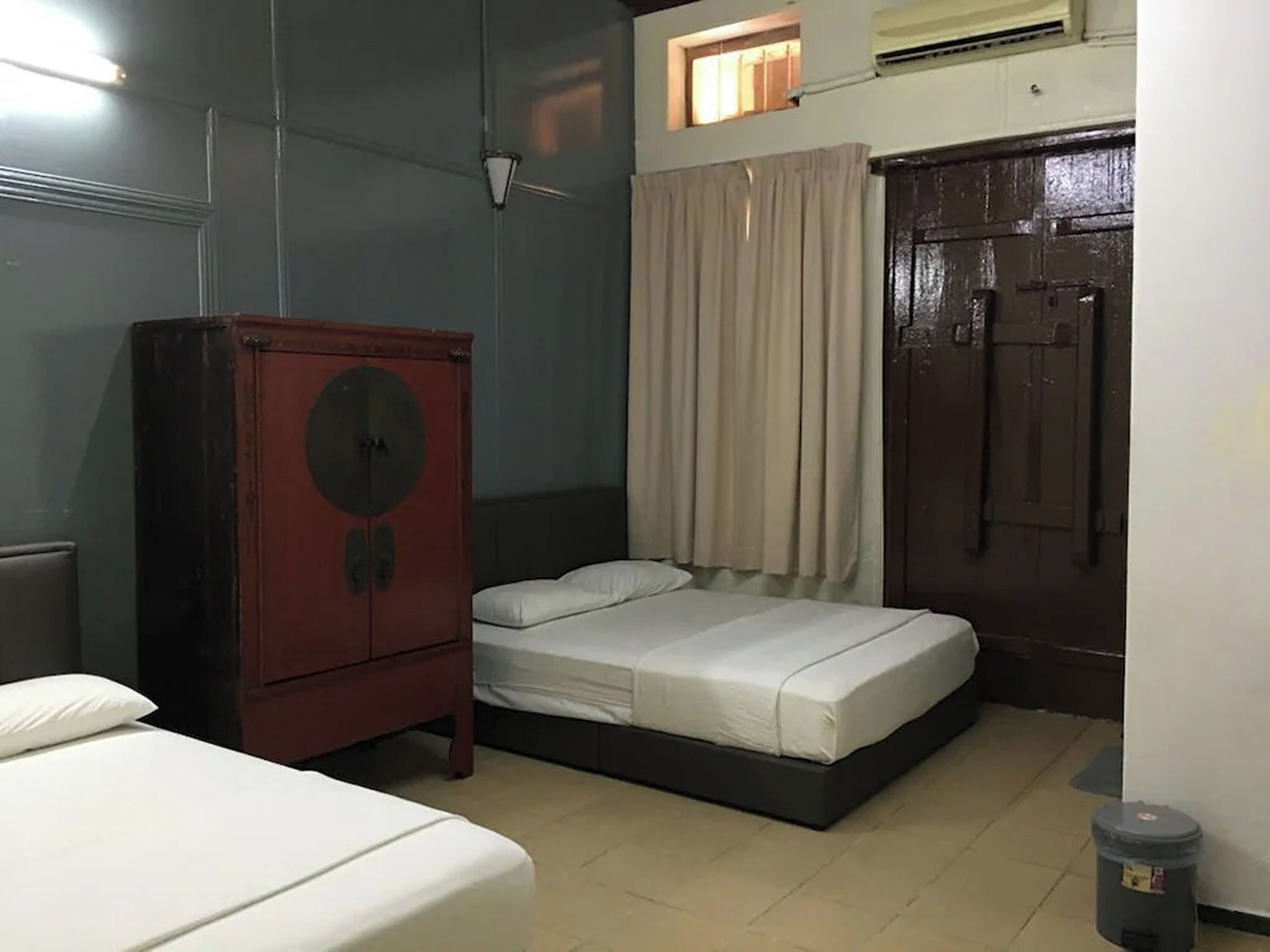 Una habitación pequeña y tenuemente iluminada, con una cama individual, un armario de madera y una ventana con cortinas.