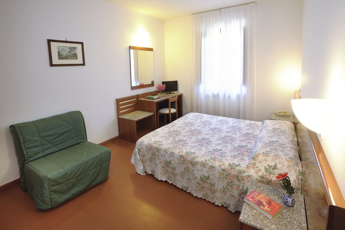 A hotel room with a green sofa, a bed with a floral bedspread, a desk with a chair, a computer, and a window with curtains.
