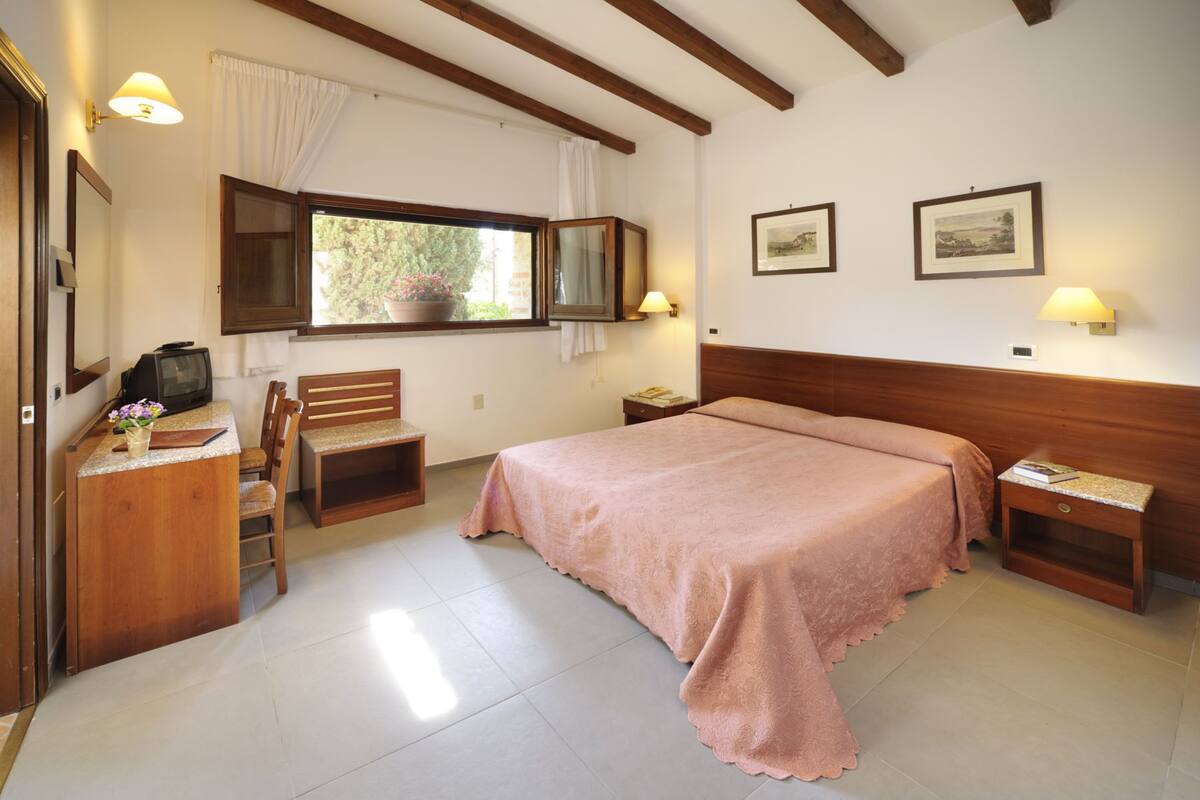 A bedroom with a large bed, wooden headboard, a desk with a television, a chair, a window with curtains, and two wall-mounted lamps.