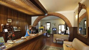 A reception area with a wooden reception desk, a receptionist, a sofa, and a large mirror. - Hotel Villalago (Magione)