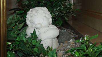 En hvid stenskulptur af en figur med rundt hoved og lukkede øjne, omgivet af grønt løv og småsten.