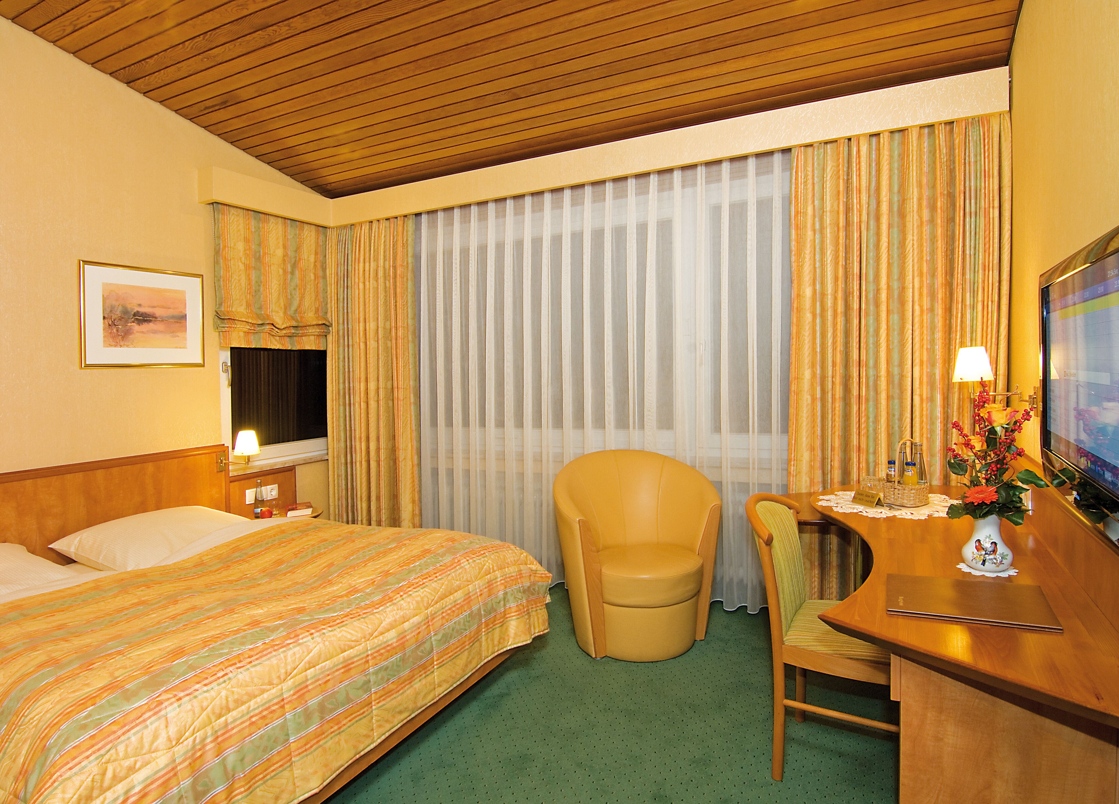 Una camera d'albergo con un letto, una scrivania con sedia, una televisione e una finestra con tende.