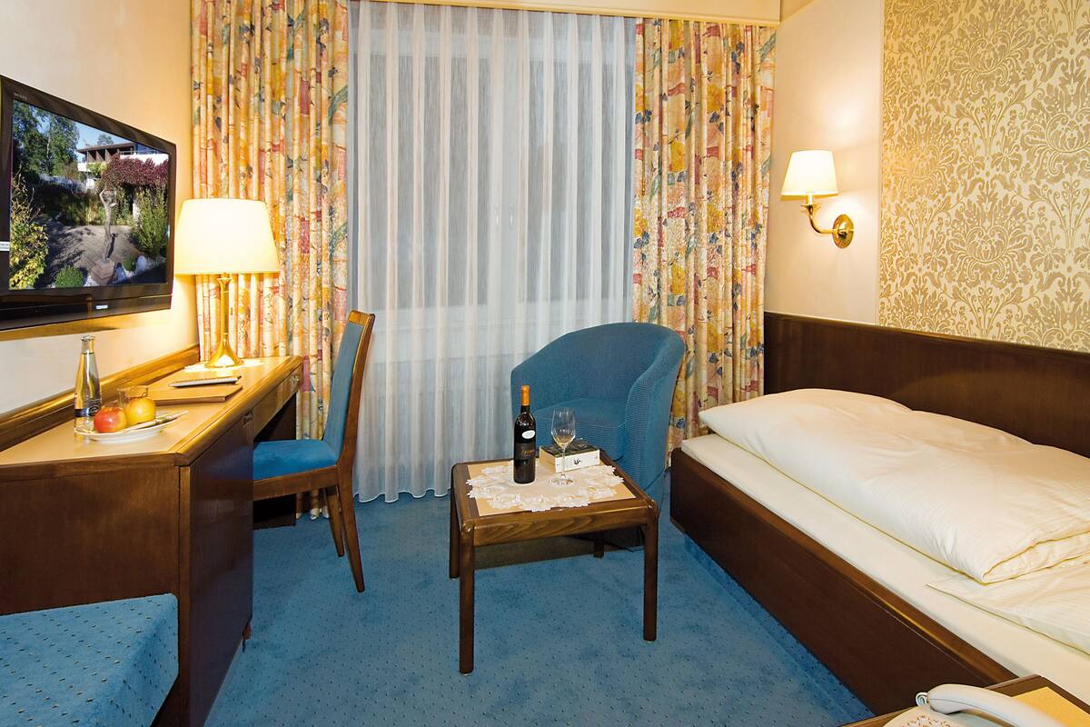 A hotel room with a bed, a desk with a chair, a television, a table with a bottle of wine and a glass, and a telephone.