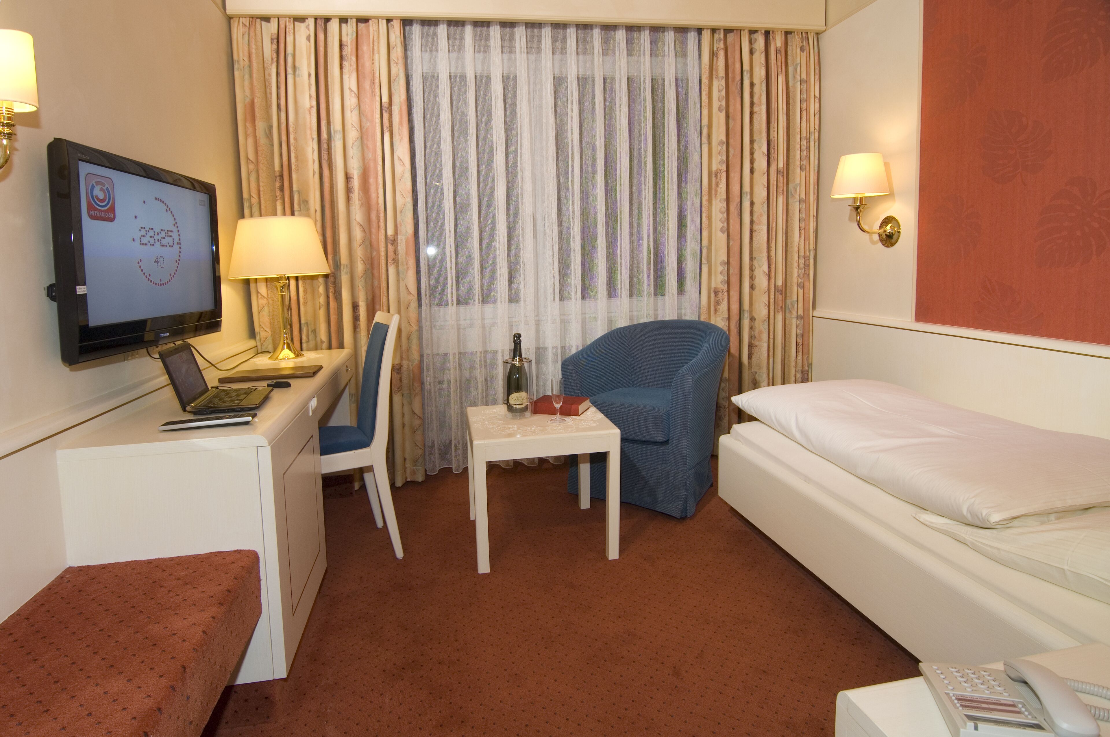 Habitación de hotel con cama, escritorio con una computadora portátil, silla, pequeña mesa con una botella de vino y vaso, y televisor en la pared.