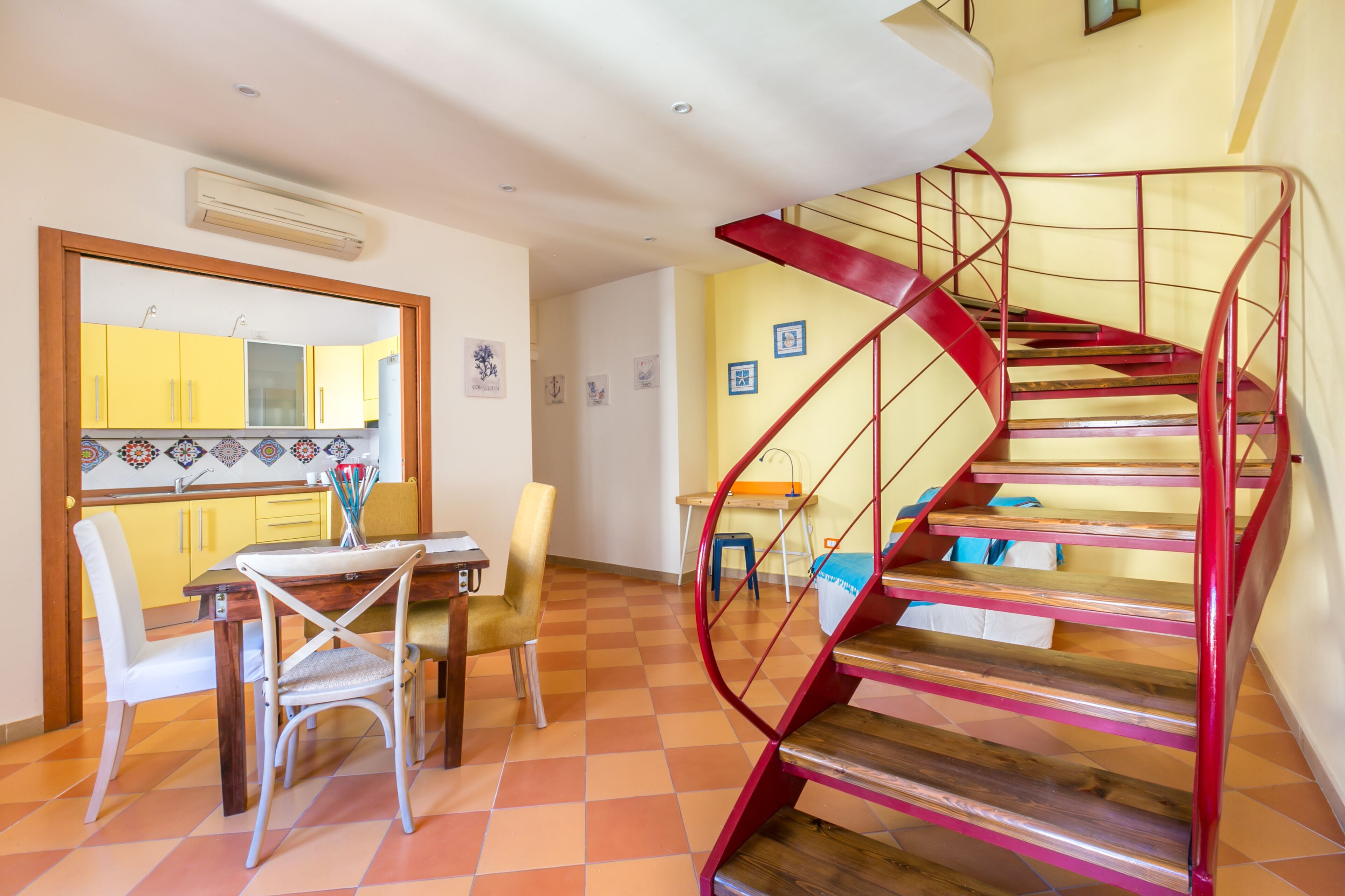 A dining area with a wooden table and chairs, a kitchen with yellow cabinets, and a red spiral staircase.