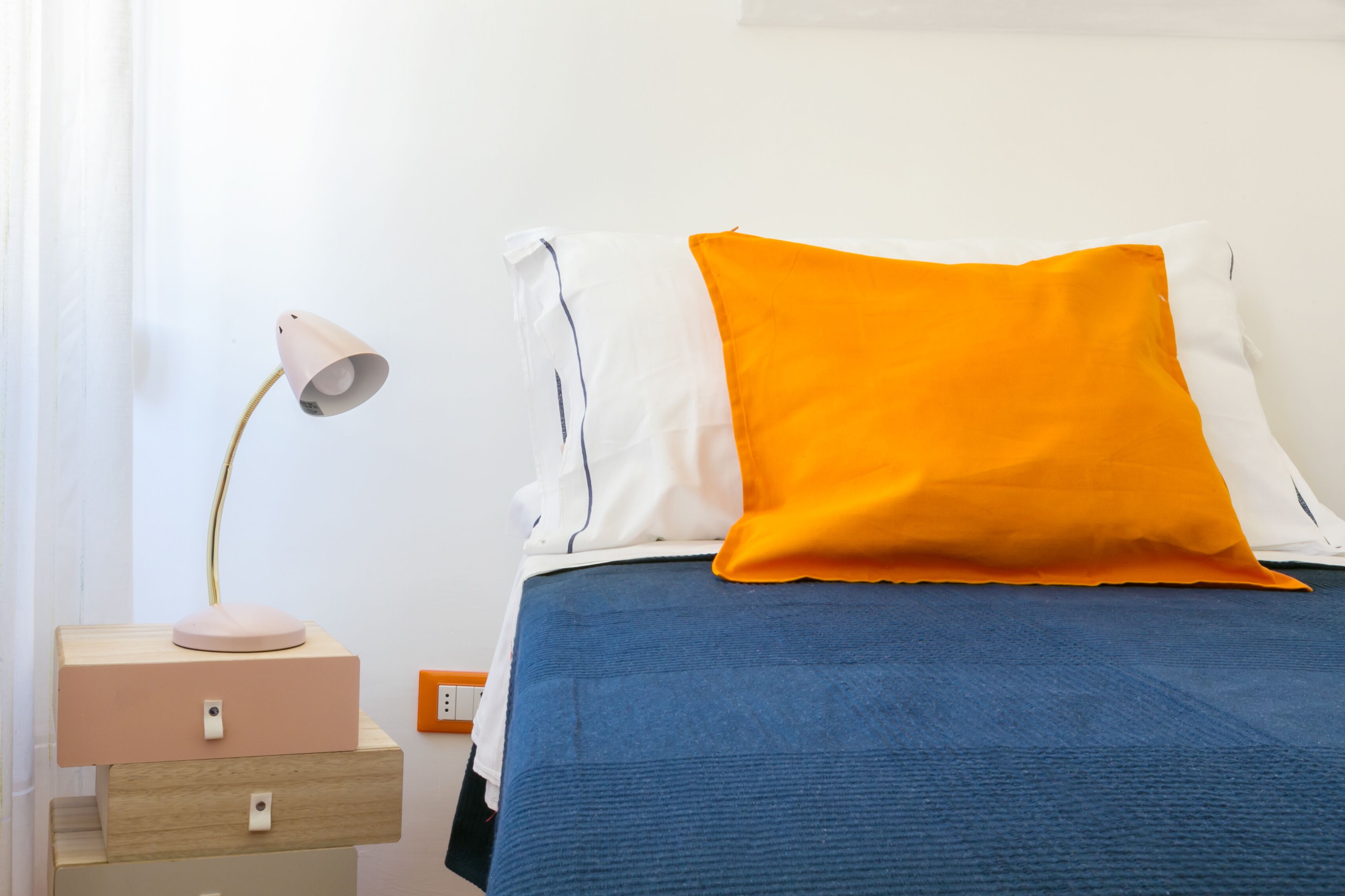A neatly made bed with a blue bedspread and two pillows, one white and one orange, in a simple room with a bedside lamp and a small wooden nightstand.
