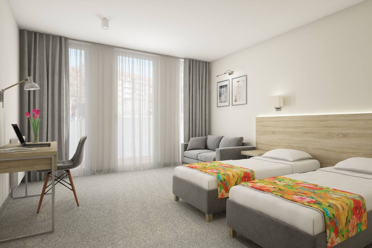 A hotel room with two beds, a desk with a laptop, a chair, and a large window with curtains.