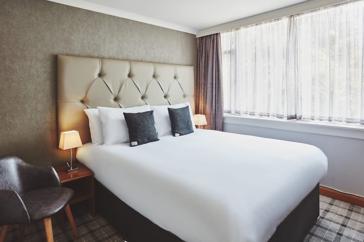A hotel room with a large bed, two pillows, a nightstand with a lamp, a chair, and a window with curtains.
