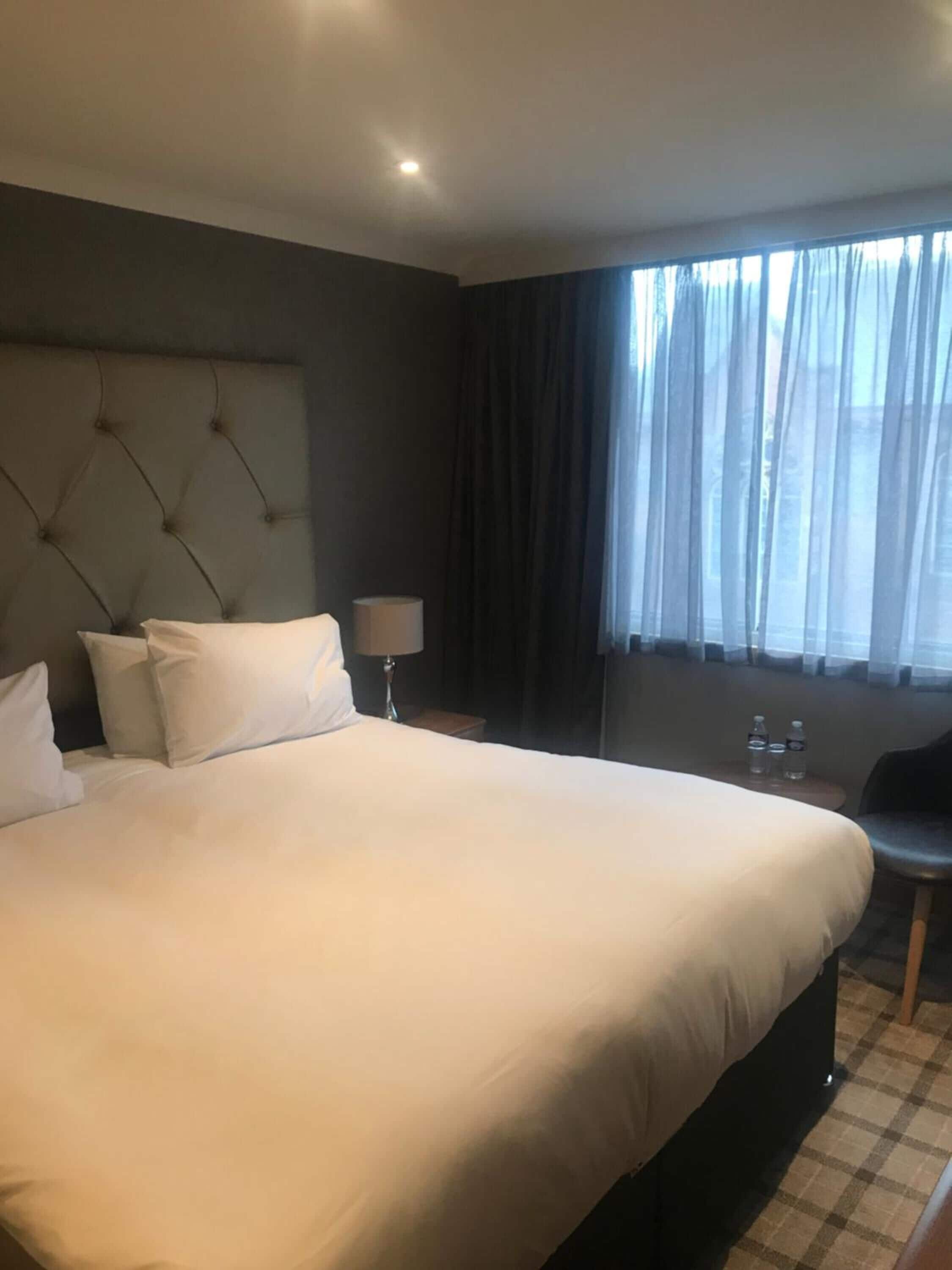 Habitación de hotel con una cama grande, cabecera, mesita de noche con lámpara, una pequeña mesa con botellas de agua y una ventana con cortinas.