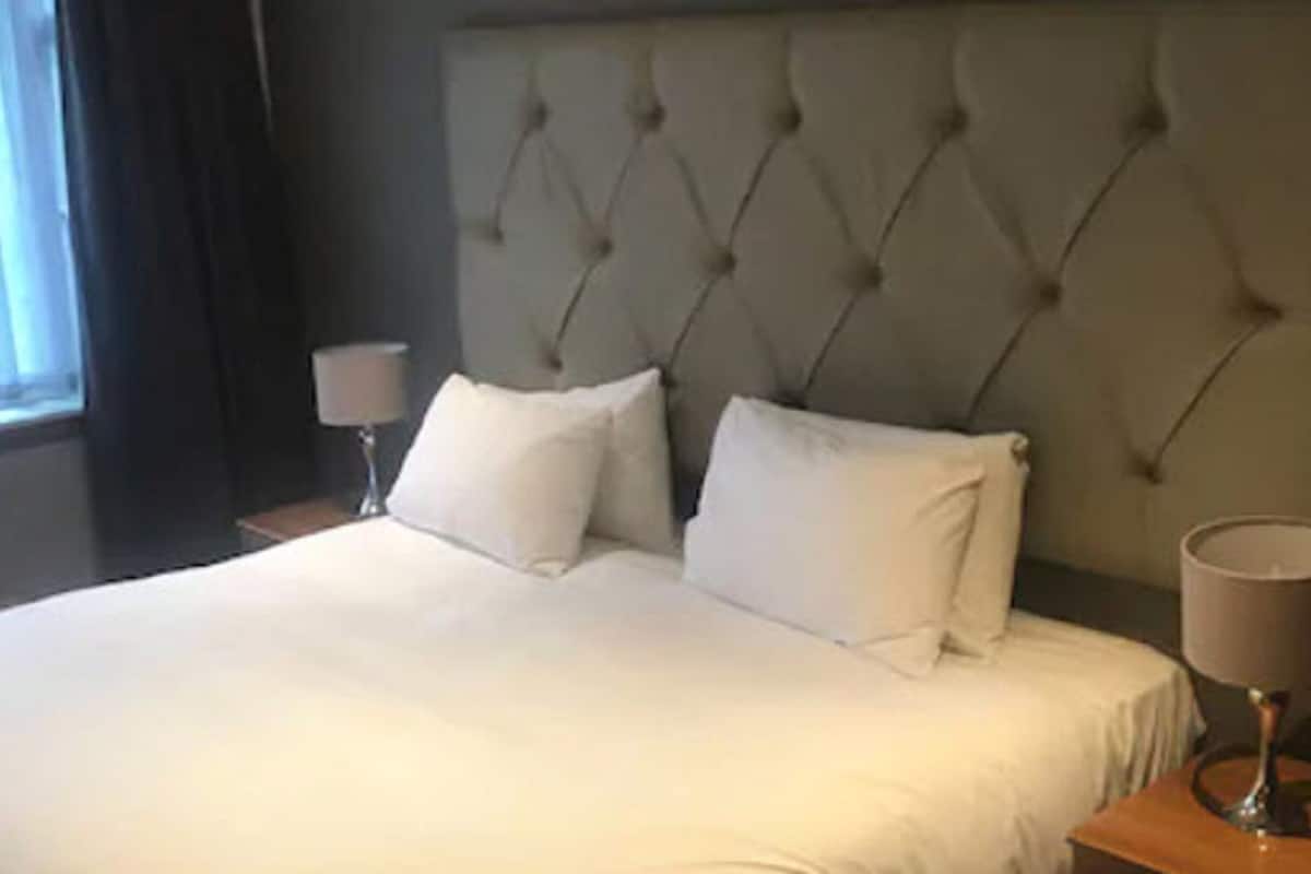 A hotel room with a large bed, two pillows, a nightstand with a lamp, and a window with curtains.