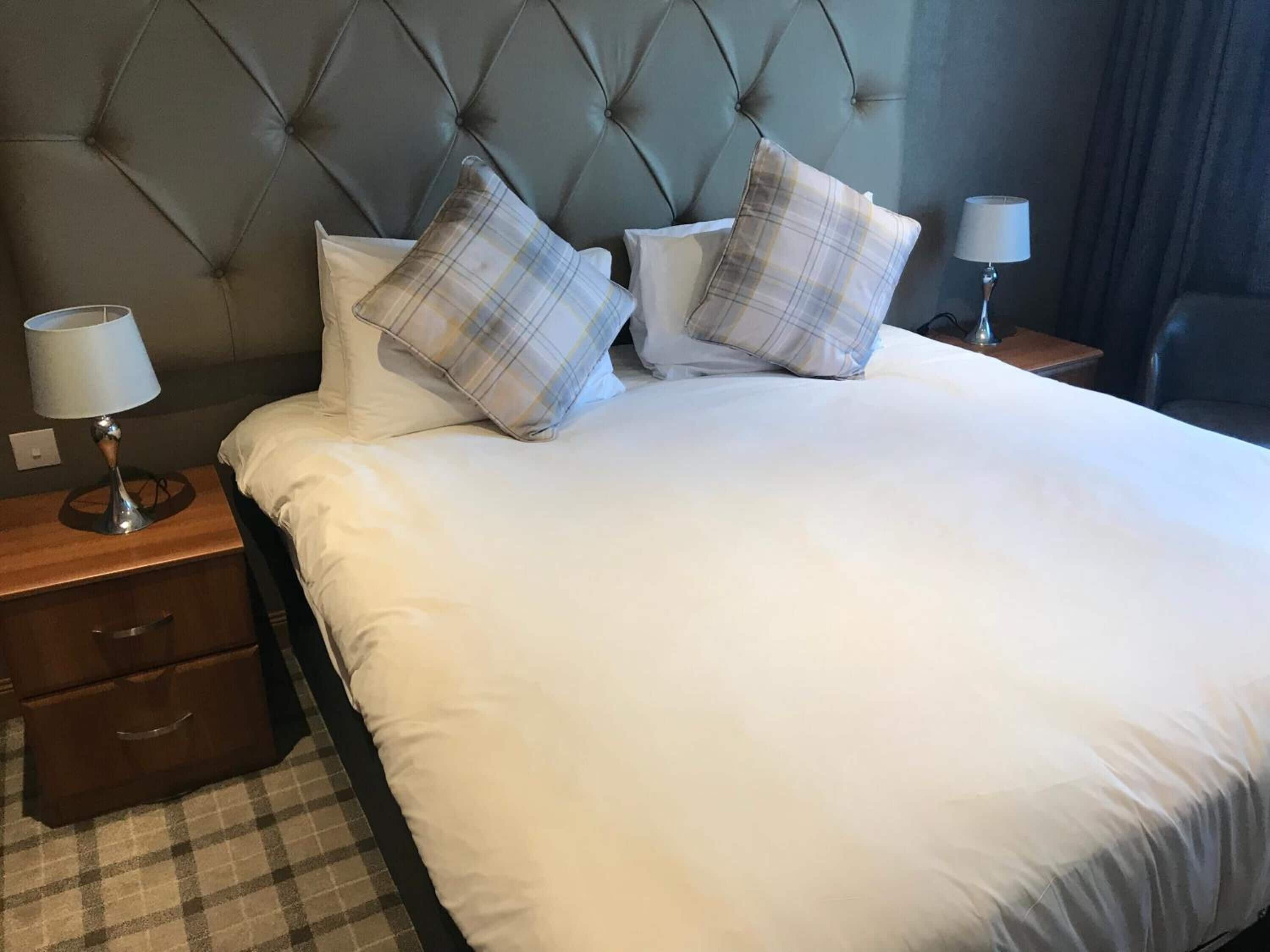 A neatly made bed with white linens and plaid pillows, flanked by two bedside tables with lamps, in a room with a tufted headboard.