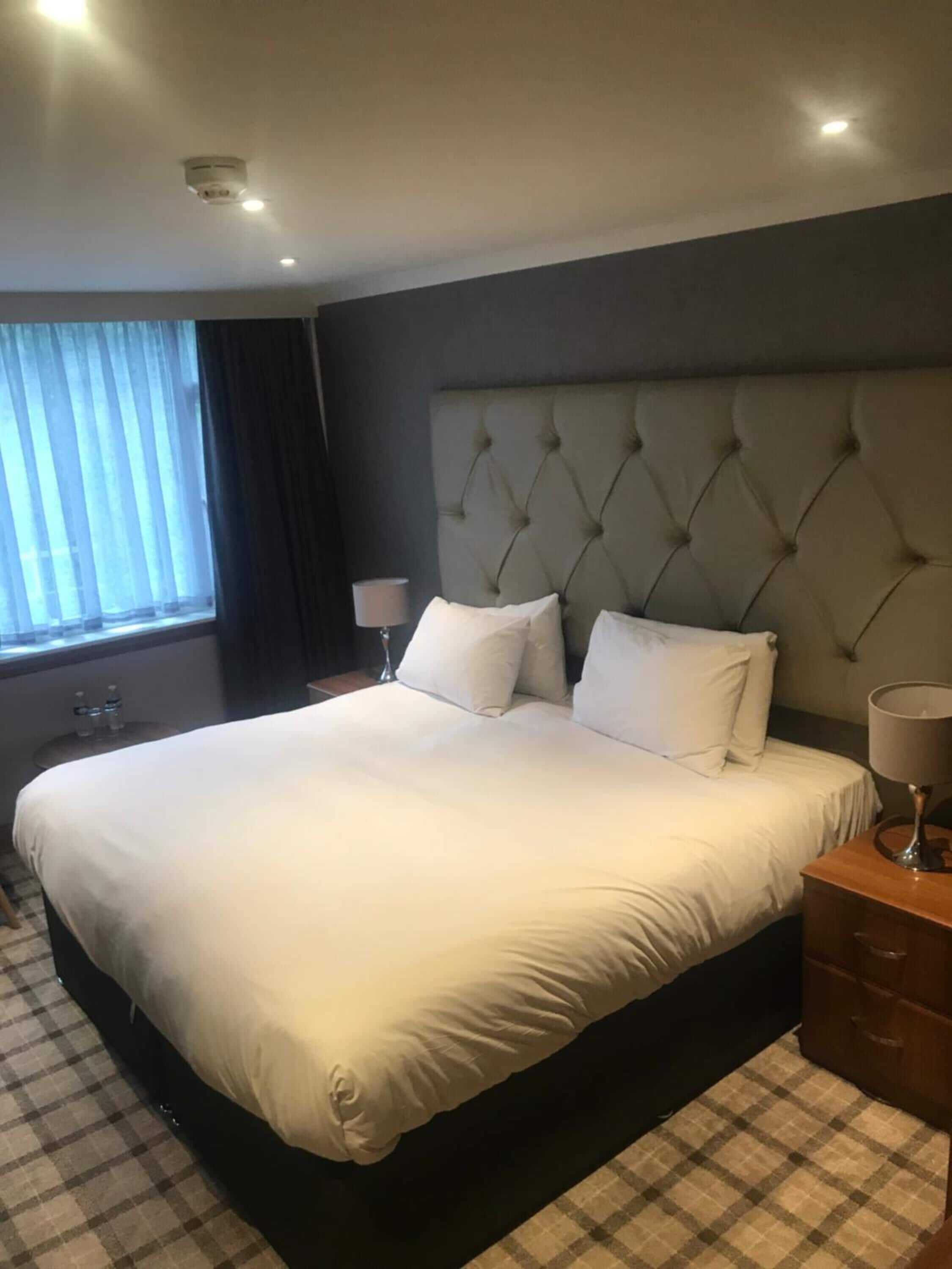 Ein Hotelzimmer mit einem großen Bett, einem mit Knöpfen besetzten Kopfteil, einem Nachttisch mit Lampe und einem Fenster mit Vorhängen.