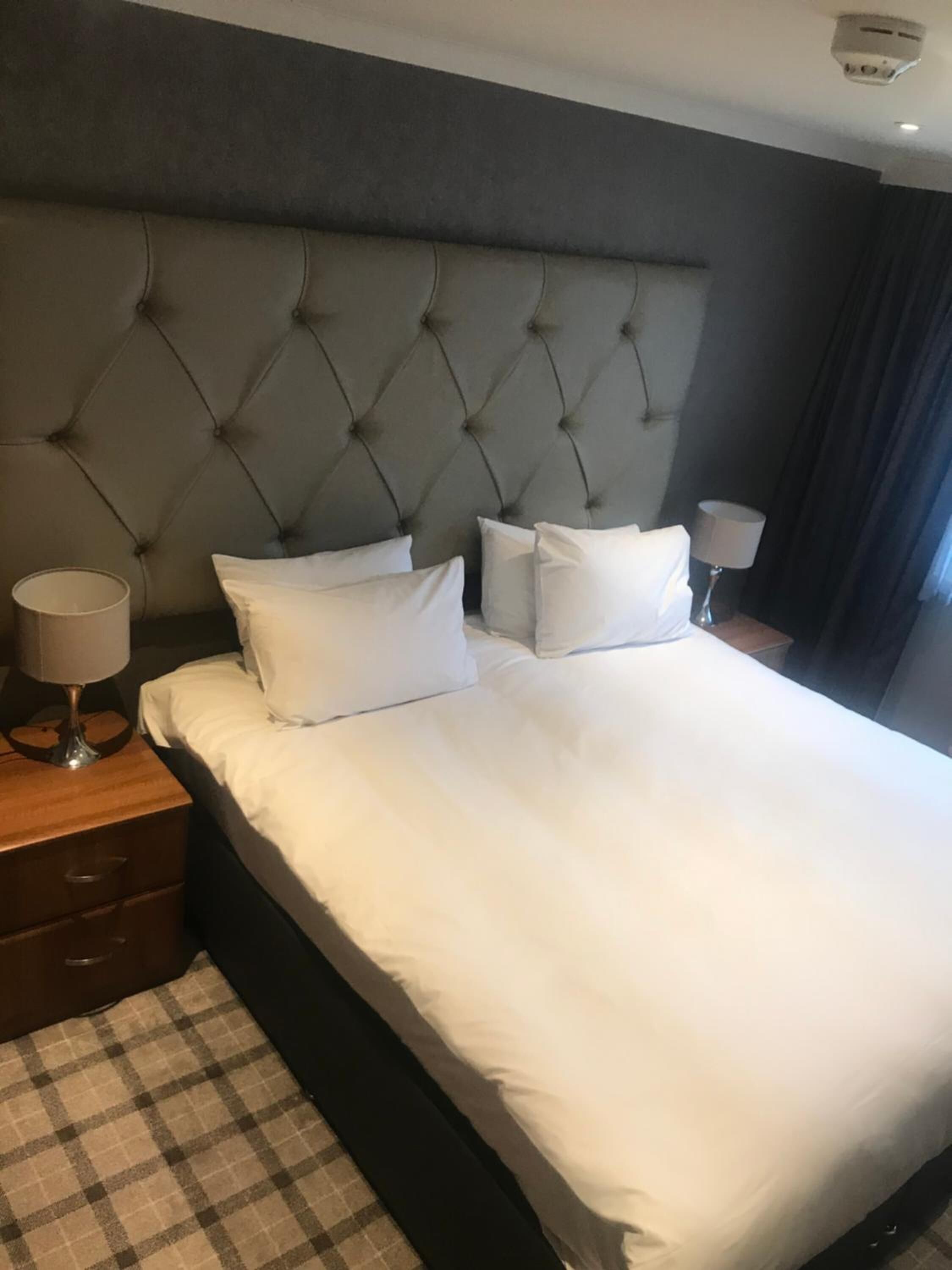 Una cama bien hecha con un cabecero capitonado, una lámpara de noche y un suelo a cuadros.