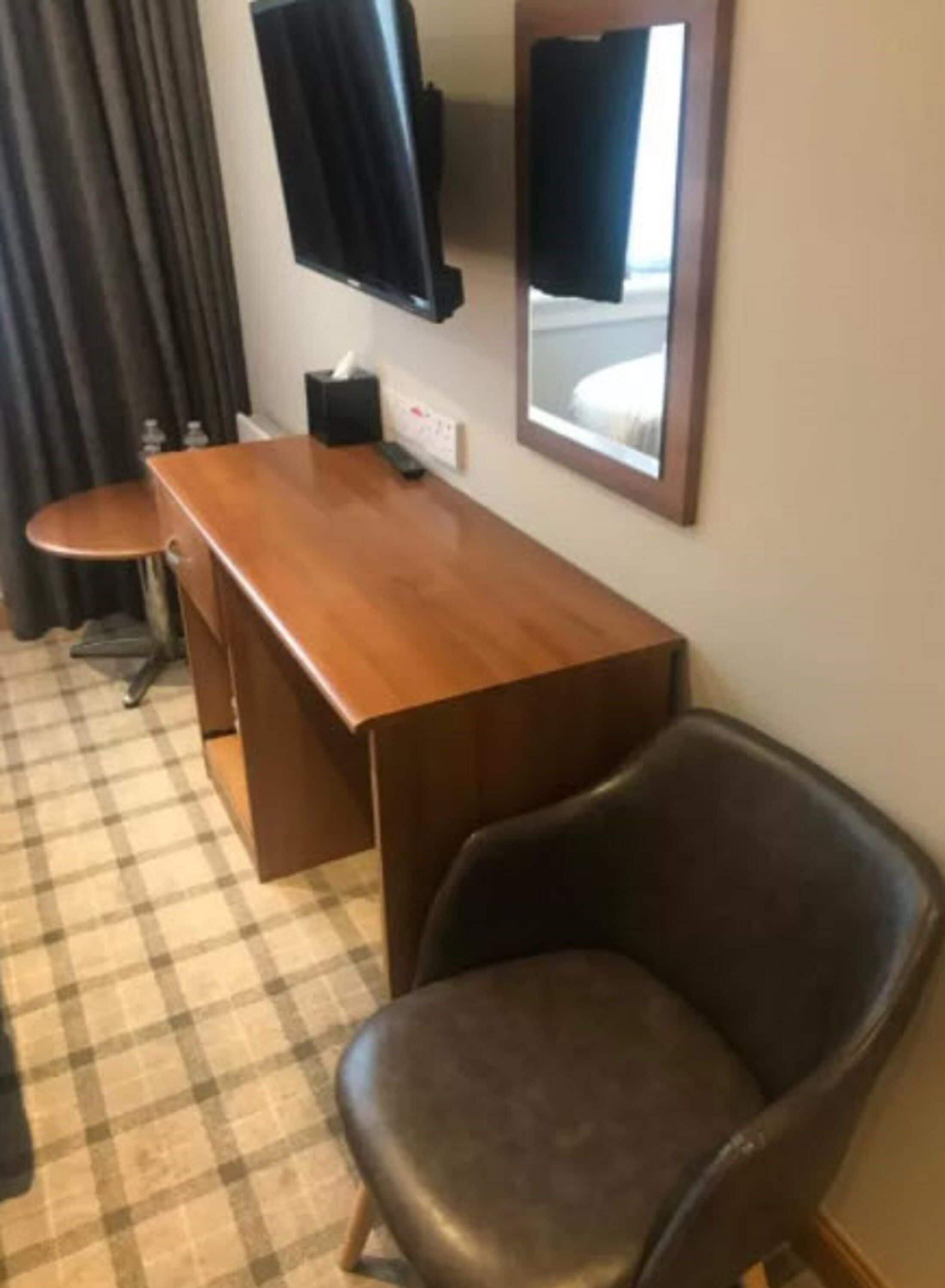 A hotel room with a wooden desk, a black leather chair, a small table, a TV mounted on the wall, and a checkered floor.