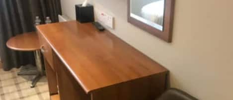 A hotel room with a wooden desk, a black leather chair, a small table, a TV mounted on the wall, and a checkered floor.