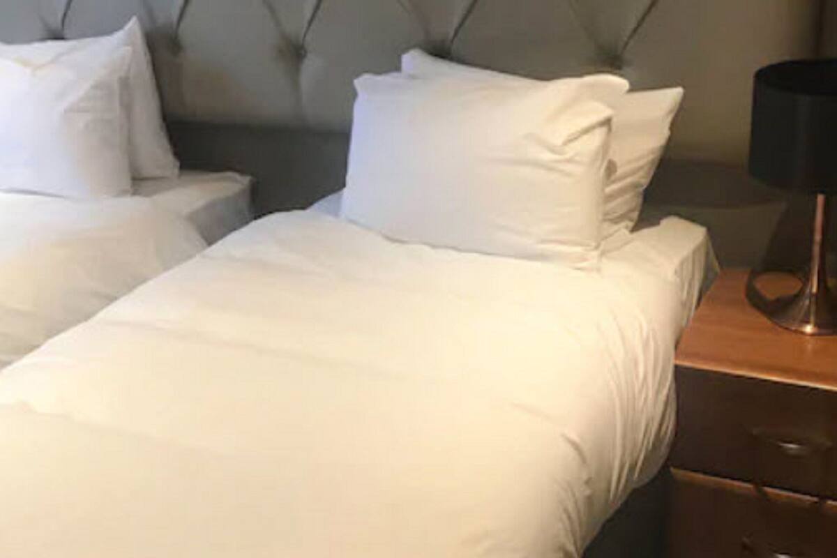 A hotel room with two single beds, each with white linens and pillows. There is a wooden nightstand with a lamp on it.