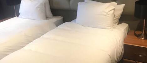 A hotel room with two single beds, each with white linens and pillows. There is a wooden nightstand with a lamp on it.