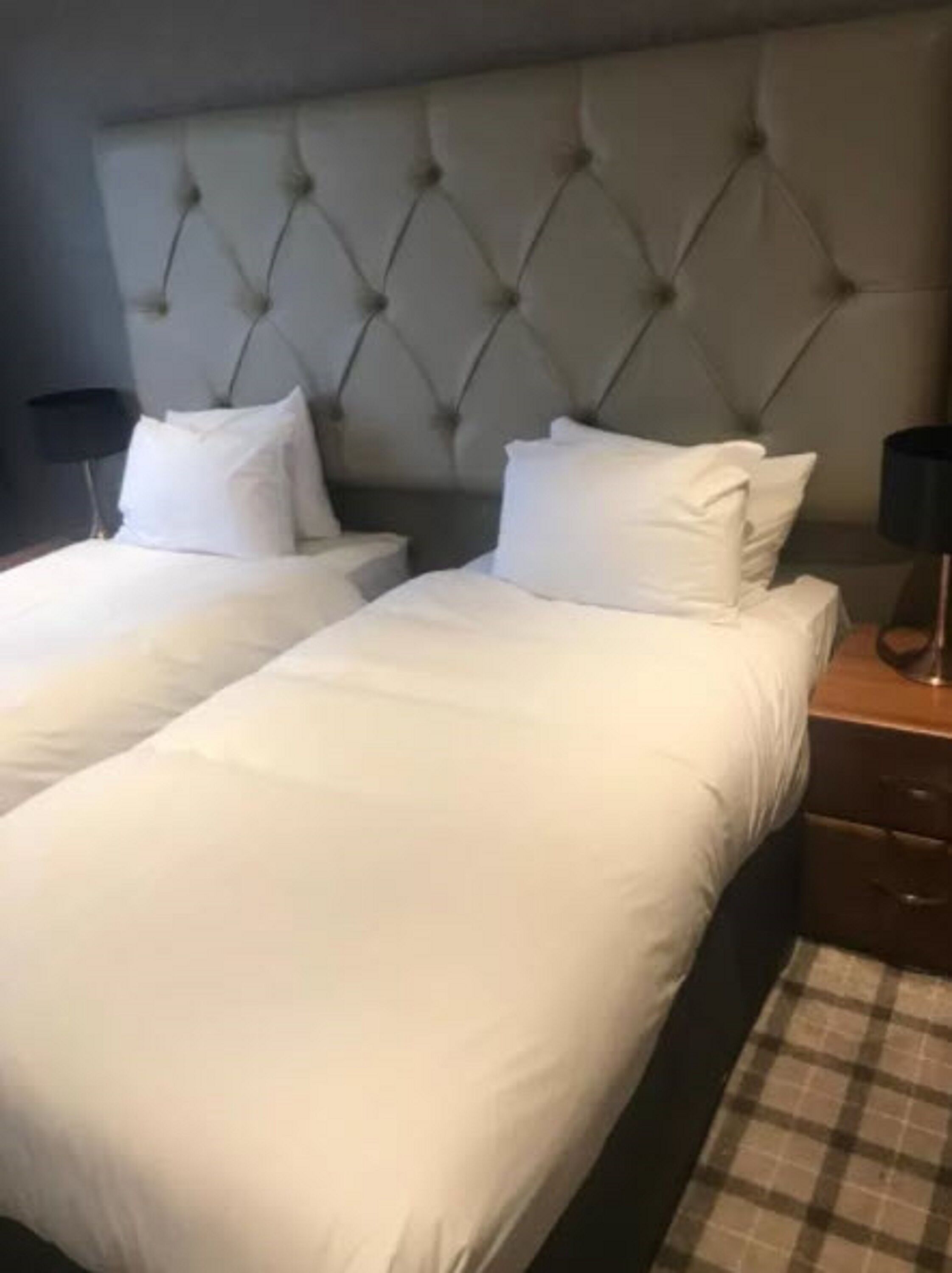 Et hotelværelse med to senge, begge med hvide sengetøj og en quiltet sengegavl. Der er to natborde med lamper og en ternet løber på gulvet.