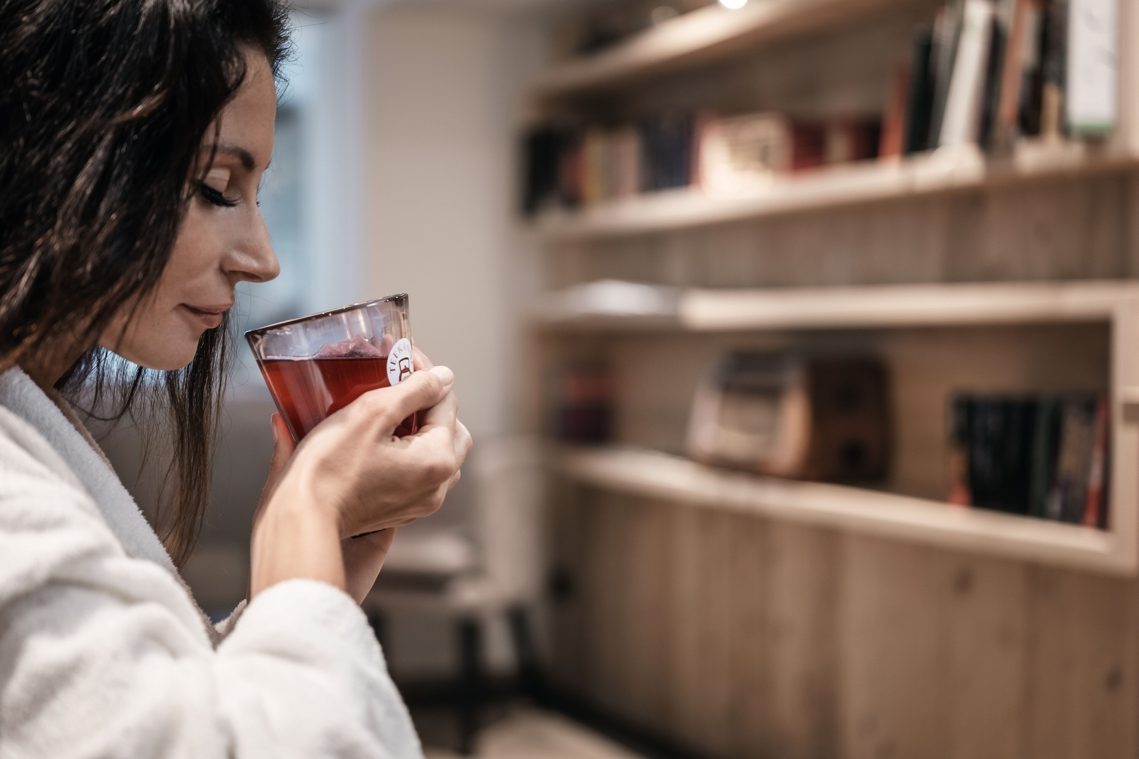 Una persona sostiene un vaso con una bebida en una habitación con estanterías de madera y libros.