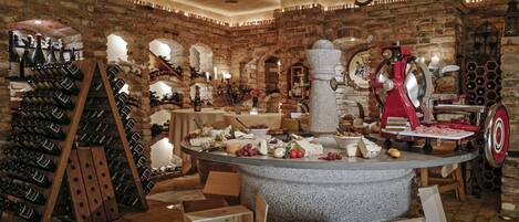 A wine cellar with wooden racks of wine bottles, a stone table with cheese and grapes, and a meat slicer.