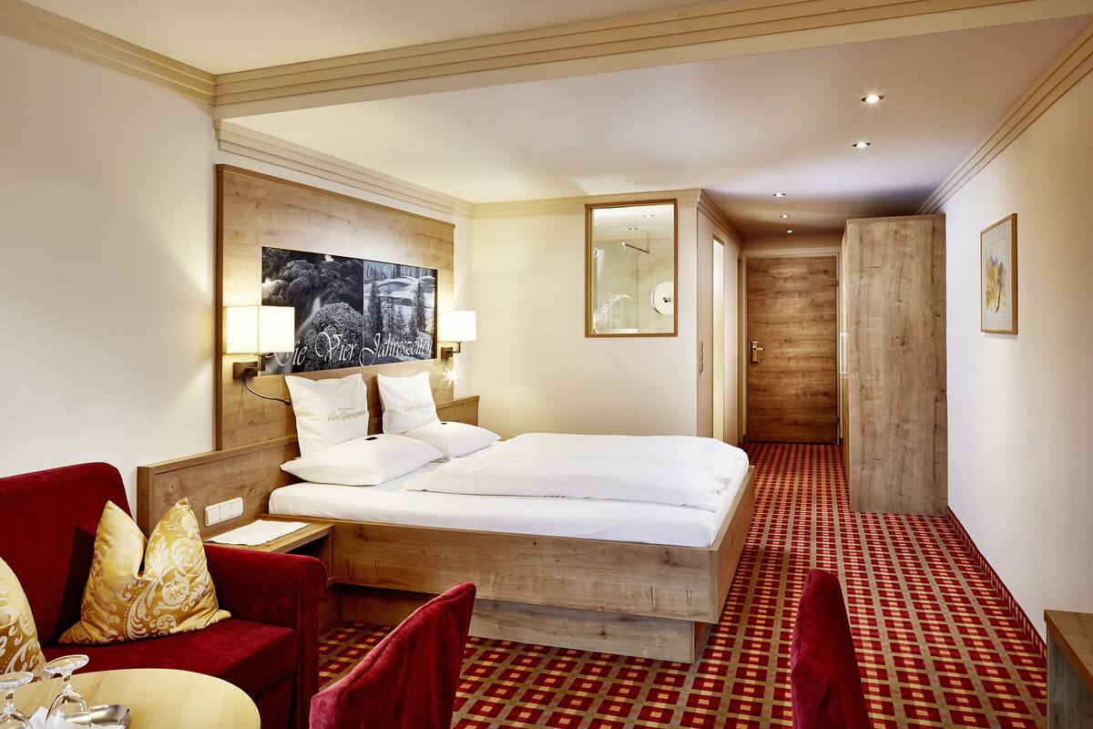 Ein Hotelzimmer mit einem großen Bett, einem roten Sessel, einem hölzernen Kopfteil, einem rot-weiß karierten Teppich und einem an der Wand befestigten Fernseher.