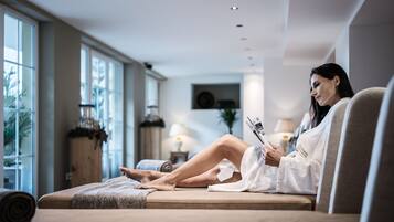 Eine Frau in einem weißen Bademantel sitzt auf einem Sofa in einem hellen Raum mit großen Fenstern, einer Lampe und einem Holzschrank.