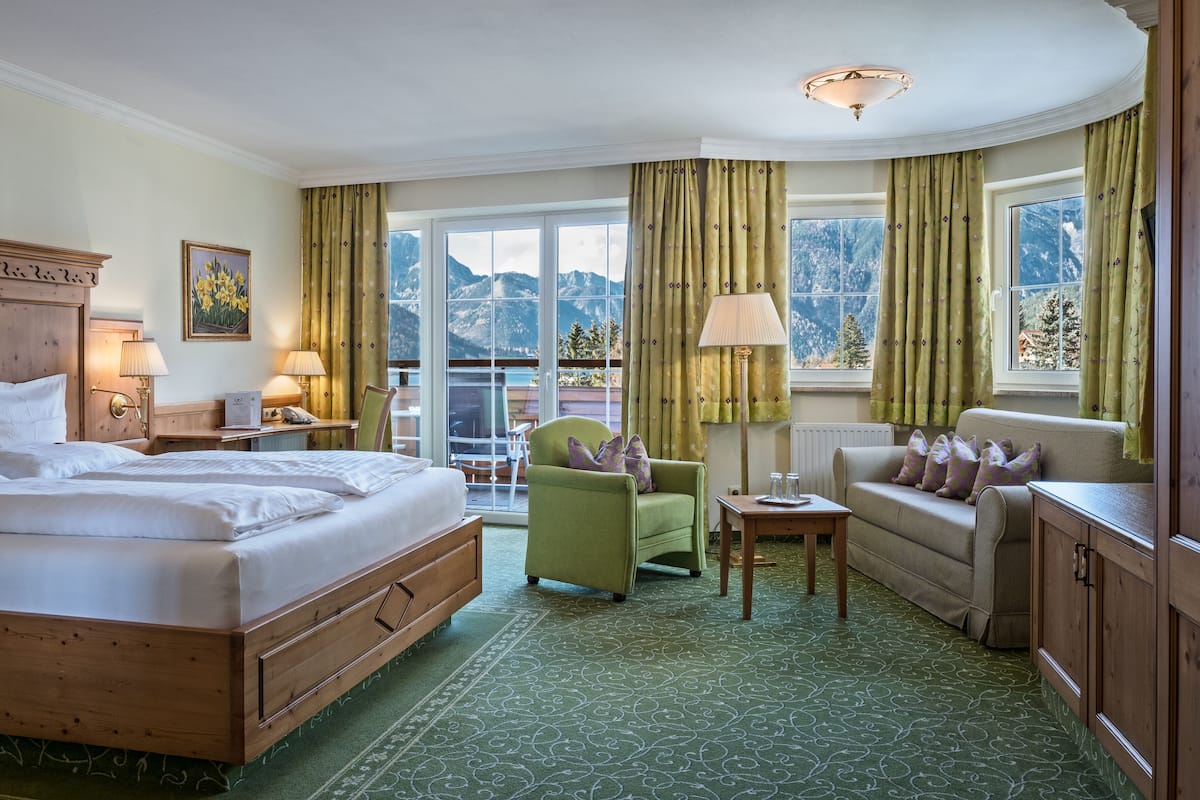 Ein Hotelzimmer mit einem großen Bett, einem grünen Sessel, einer Couch und Blick auf Berge durch die Fenster.