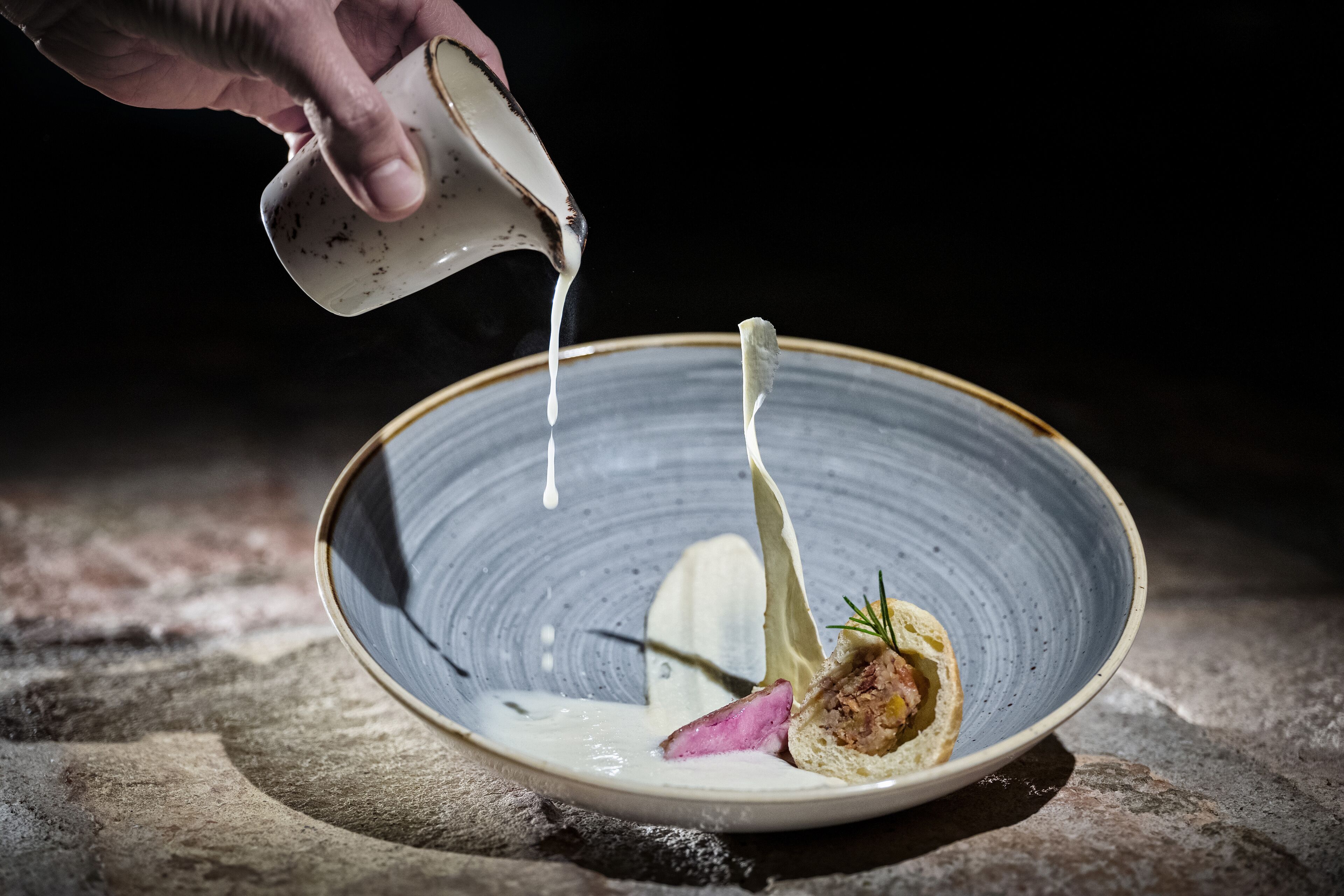 A hand pouring a white liquid into a ceramic plate with a food item and garnish.