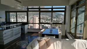 Ein modernes Wohnzimmer mit einem weißen Sofa, einem Glastisch und Blick auf eine Stadtlandschaft durch große Fenster.